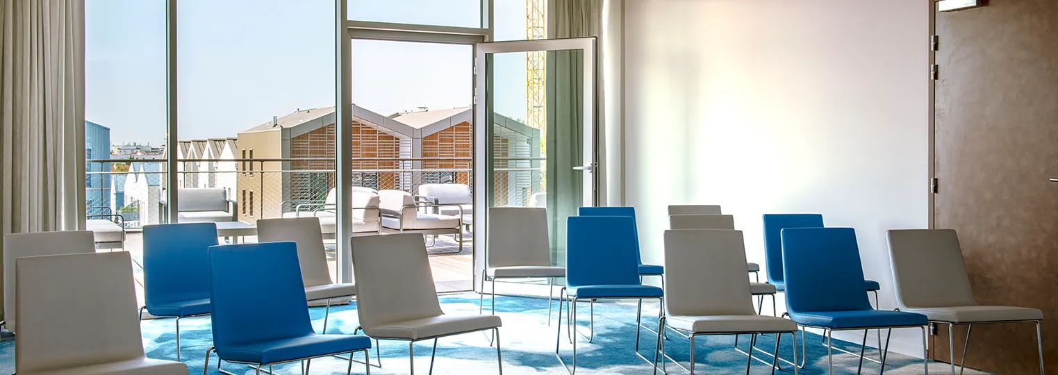 Modern hotel meeting room with blue and grey chairs arranged in rows, floor-to-ceiling windows overlooking a Bordeaux rooftop terrace with loungers, and contemporary interior design with natural light.