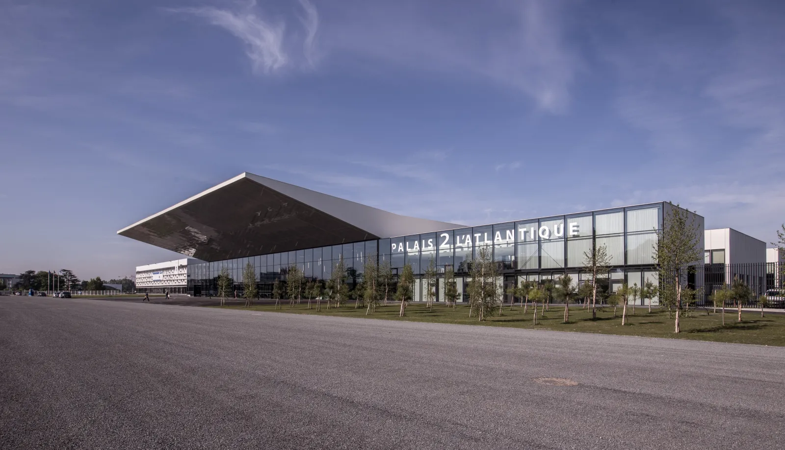 Palais 2 l'Atlantique, a modern conference and events venue in Bordeaux, featuring a large glass and steel structure with an overhanging roof, surrounded by young trees and a spacious plaza under a blue sky.