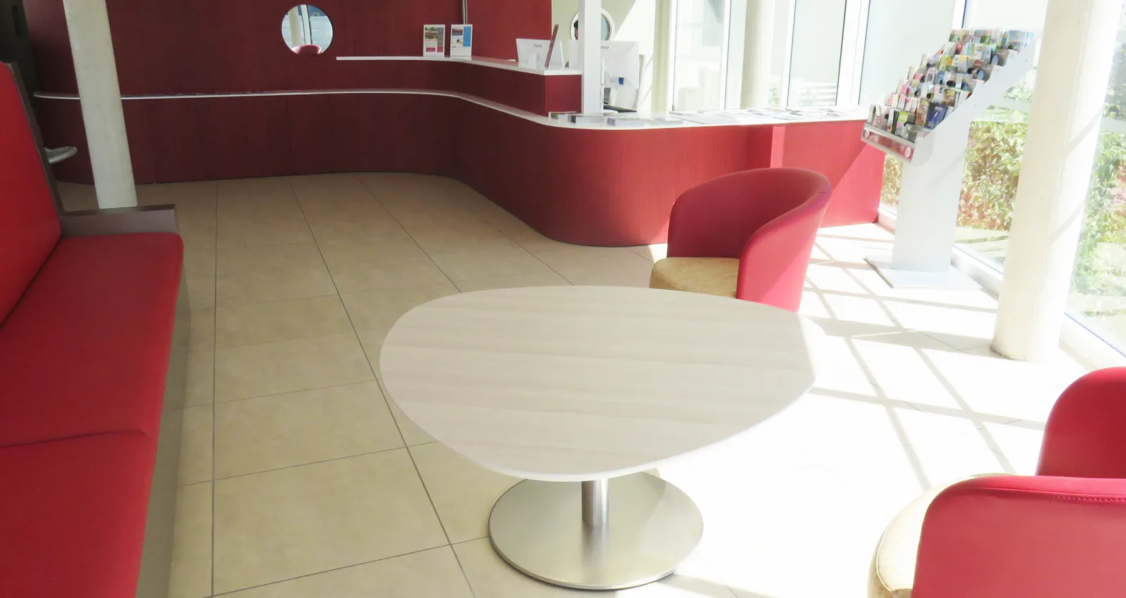 Modern reception area with red curved walls, white round table with metal pedestal base, red upholstered chairs, and bright natural light from large windows overlooking greenery.