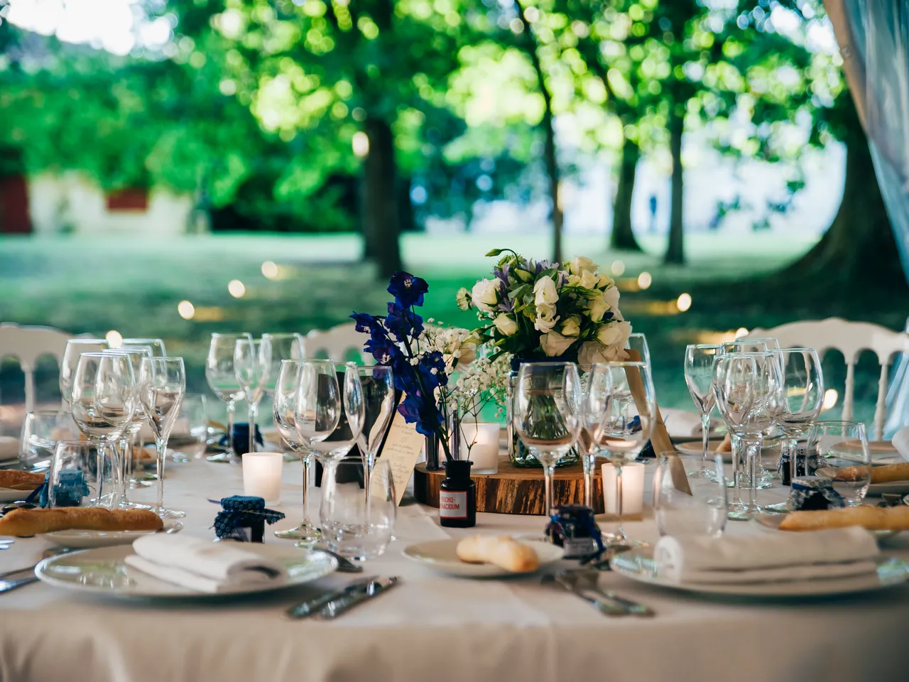 Elegant outdoor wedding reception table set with white linens, fine glassware, place settings, and floral centerpieces featuring white flowers and blue accents, with tree-lined garden backdrop