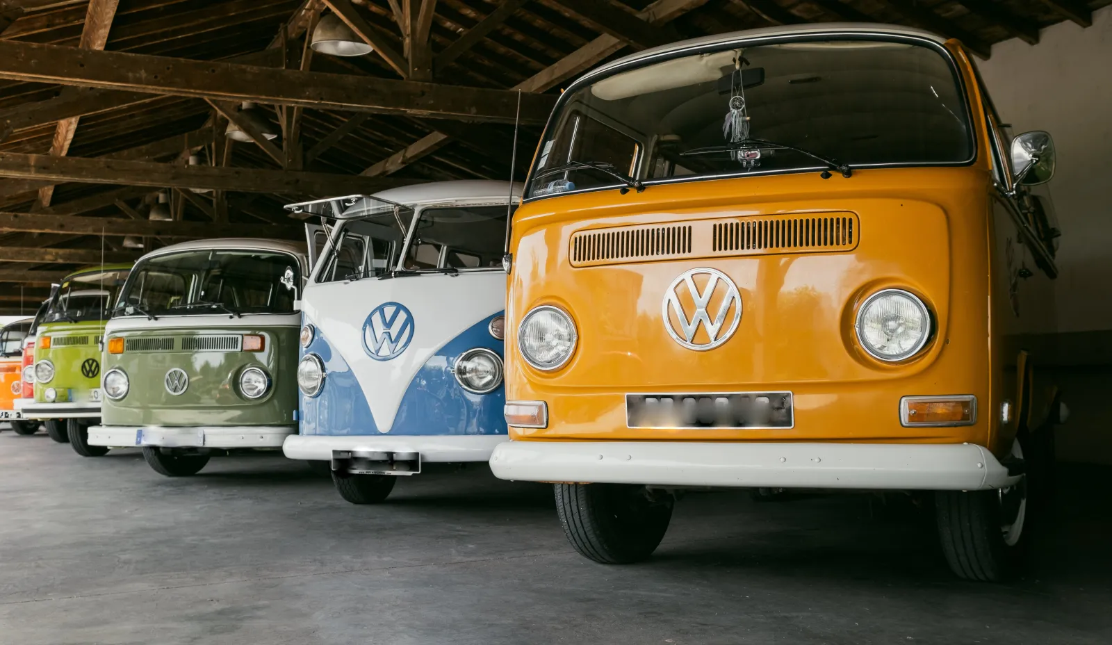 Collection of vintage Volkswagen camper vans parked in a row inside a warehouse, featuring a lime green van on the left, a blue and white van in the center, and an orange van on the right.