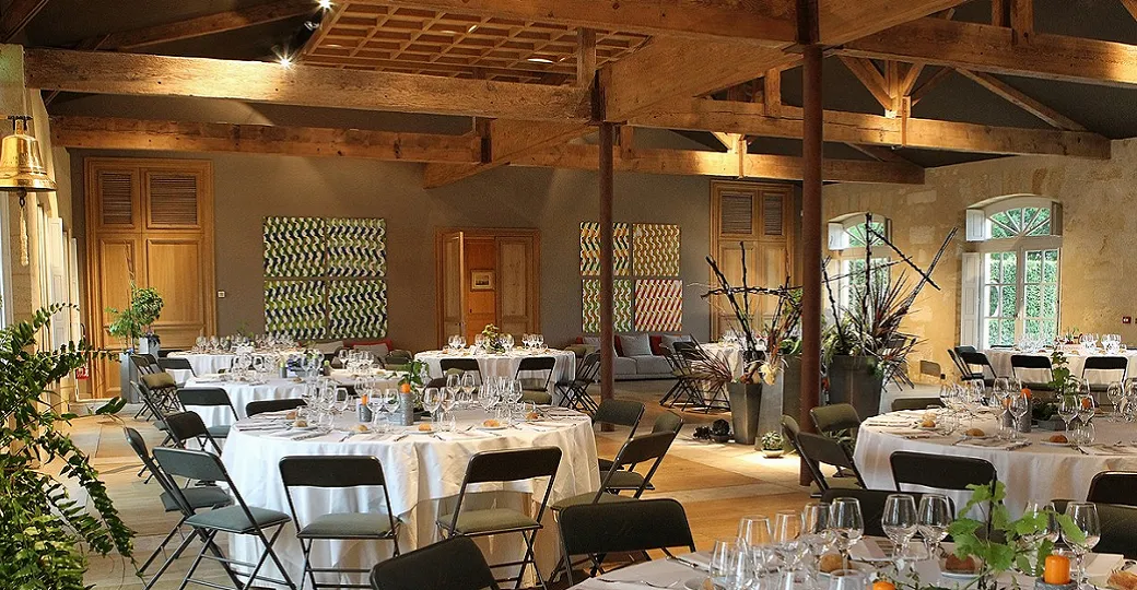 Elegant dining hall at Château Kirwan in Bordeaux with round tables set with white tablecloths, black chairs, wooden beams, and large windows overlooking the grounds.