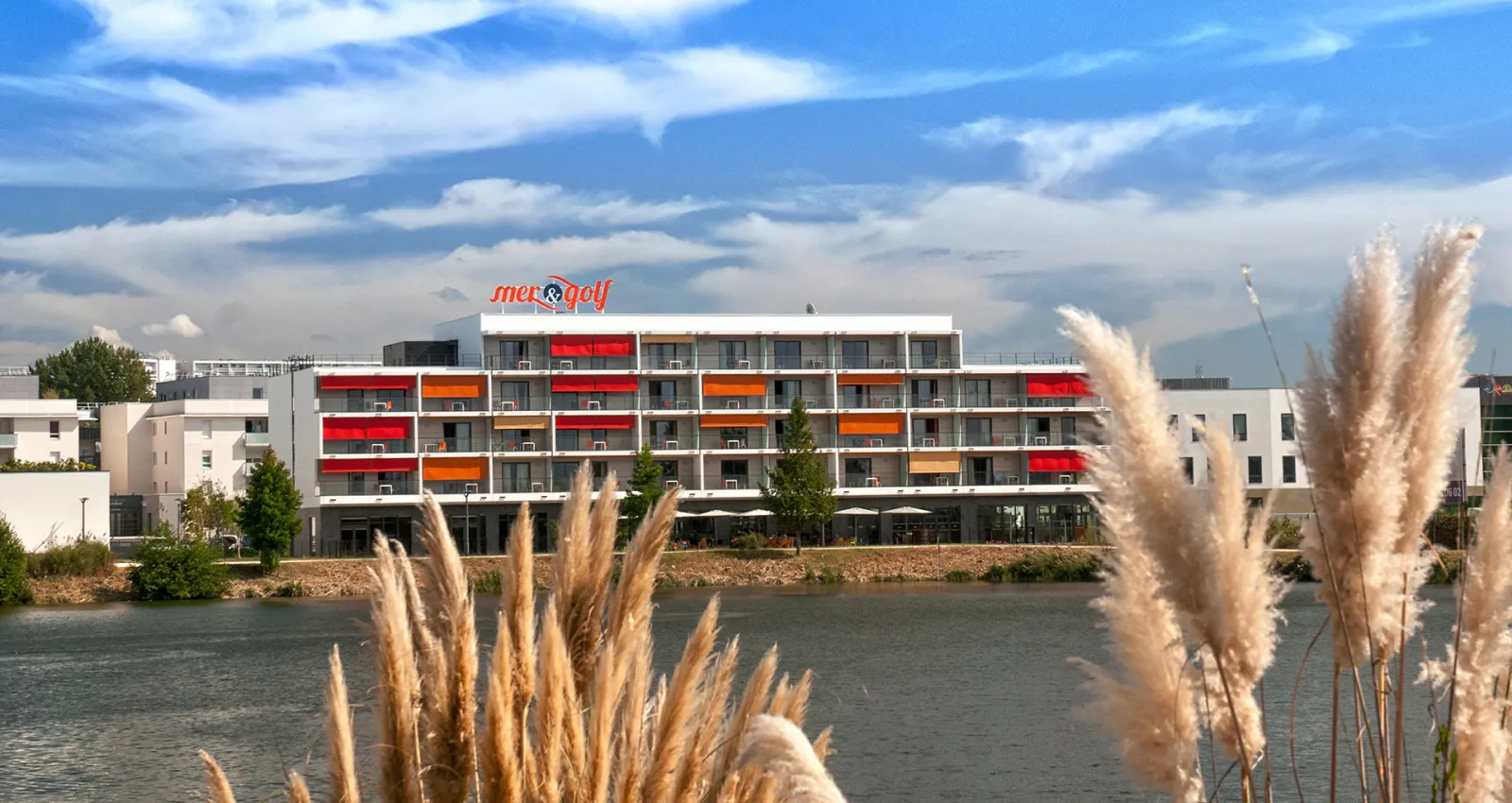Modern apartment-hotel building with red and orange balconies situated along a waterway, featuring the Mer & Golf signage on the roof, surrounded by contemporary architecture and natural vegetation.