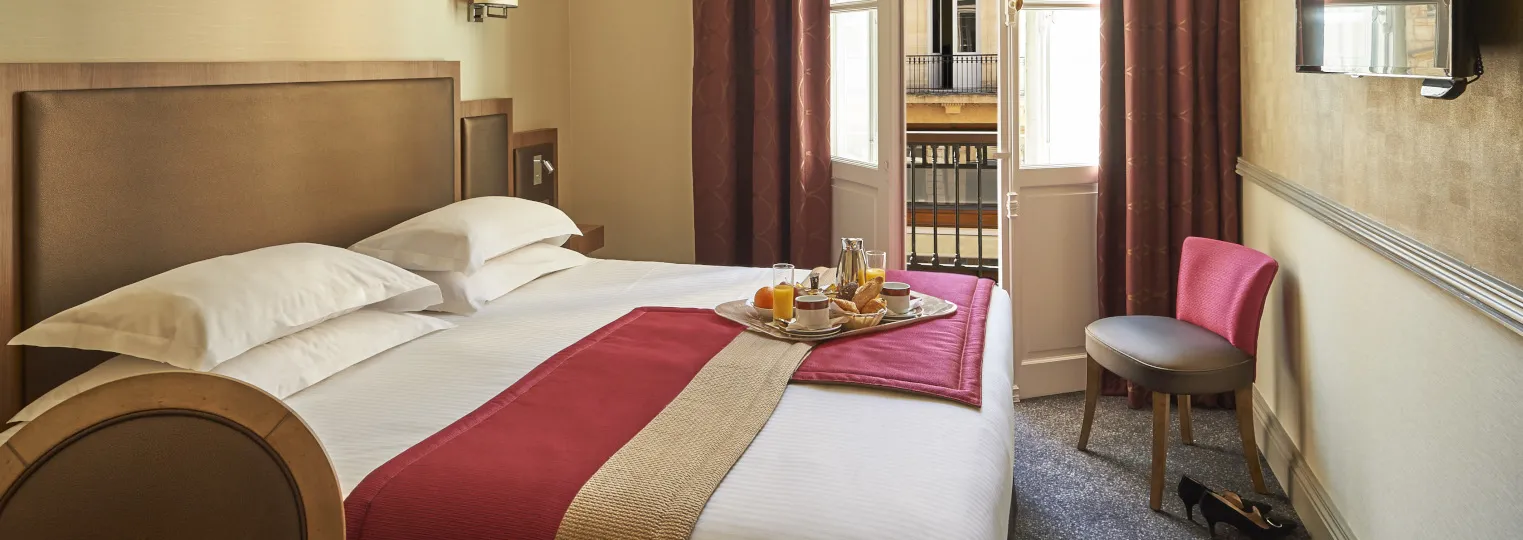 Elegant hotel bedroom at Best Western Premier Hôtel Bayonne Etche Ona in Bordeaux, featuring a king bed with white linens, red and gold bed runner, welcome tray with beverages, beige headboard, pink accent chair, burgundy curtains, and glass doors leading