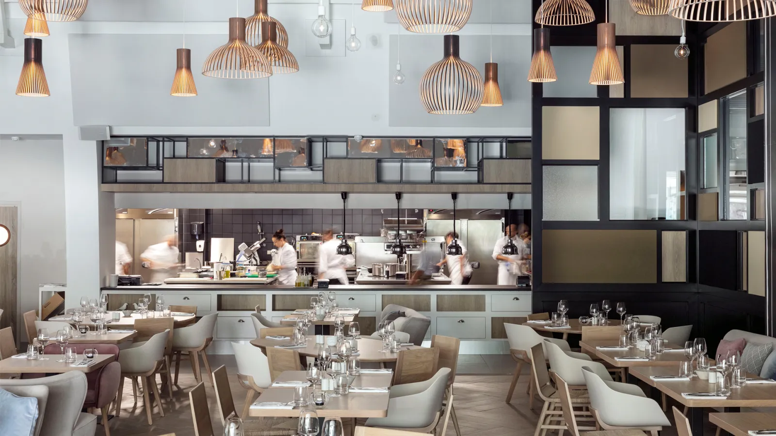 Modern restaurant interior with open kitchen concept, featuring wood and cream-colored furniture, pendant lighting, and staff in white uniforms working in the kitchen behind a glass partition.