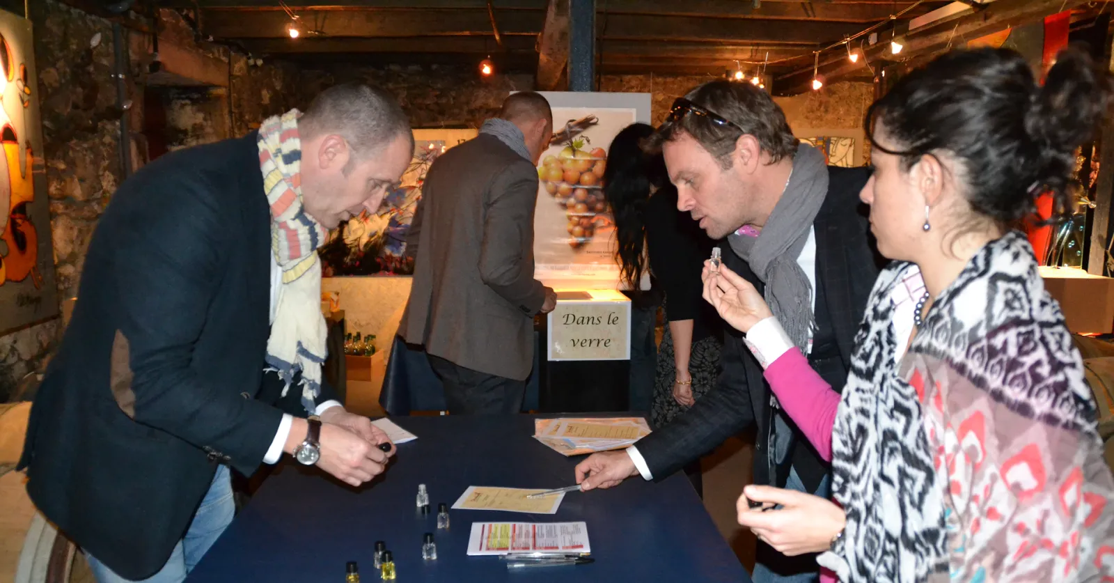 People gathered around a table at a wine tasting event in Bordeaux, examining wine glasses and promotional materials with 'Dans le verre' signage visible in an underground cellar setting with ambient lighting.