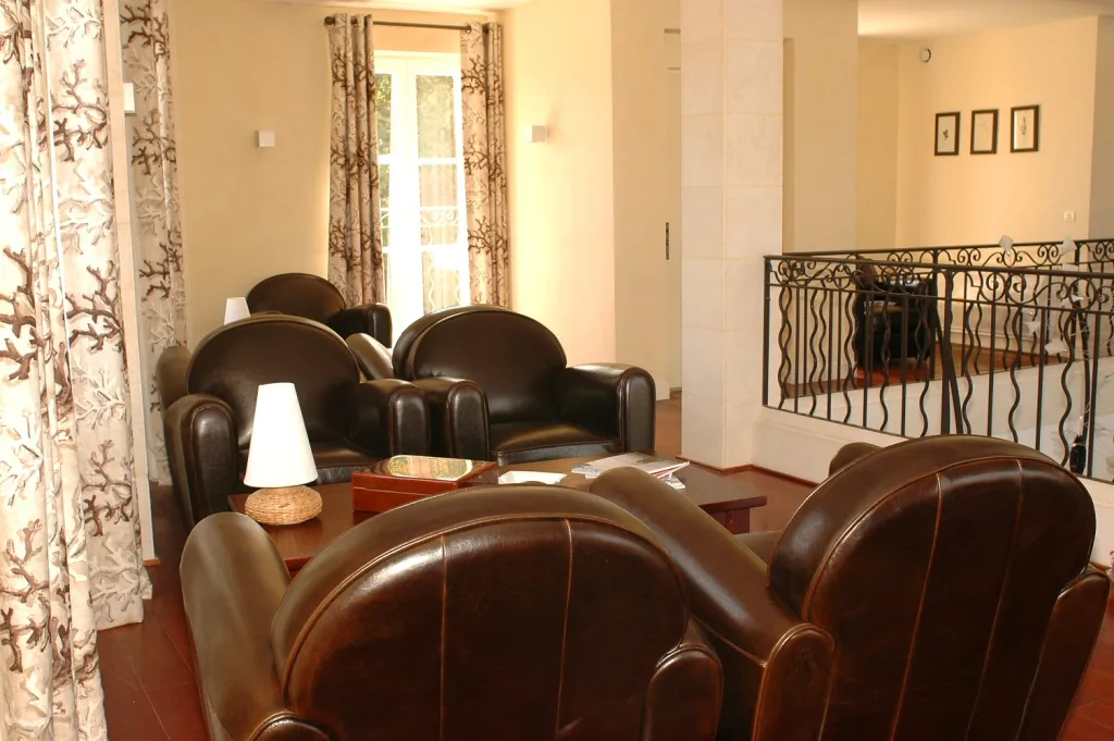 Elegant lounge area at Château Léoville Poyferré with brown leather armchairs arranged around a wooden table, featuring decorative curtains, a staircase with ornate railing, and warm beige walls.