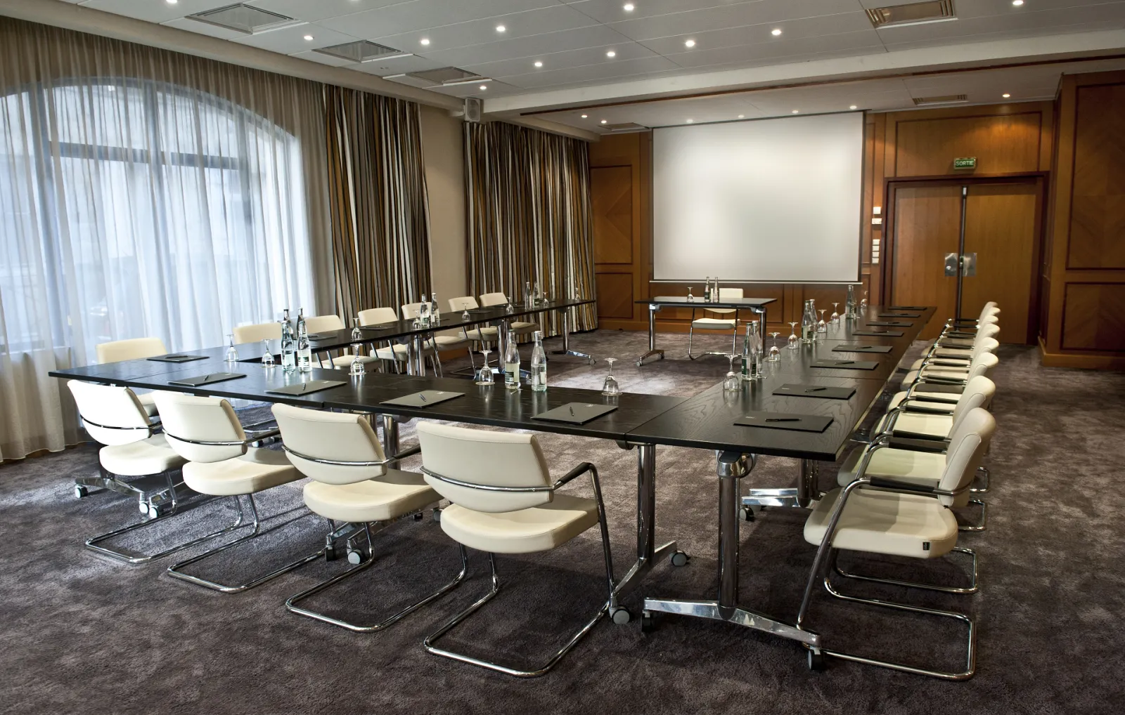 Modern conference room at Burdigala Hotel in Bordeaux, featuring a long black table with cream-colored chairs, projection screen, wooden accents, and floor-to-ceiling windows with curtains.
