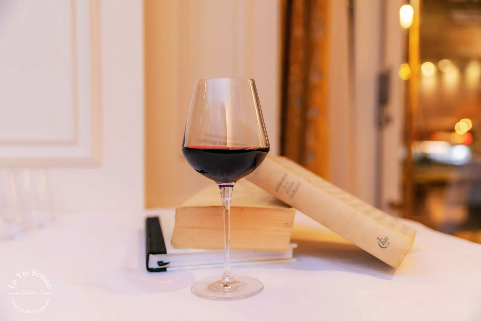 A glass of red wine displayed on a white surface next to a wine bottle with a cream-colored label, set in an elegant interior with warm lighting