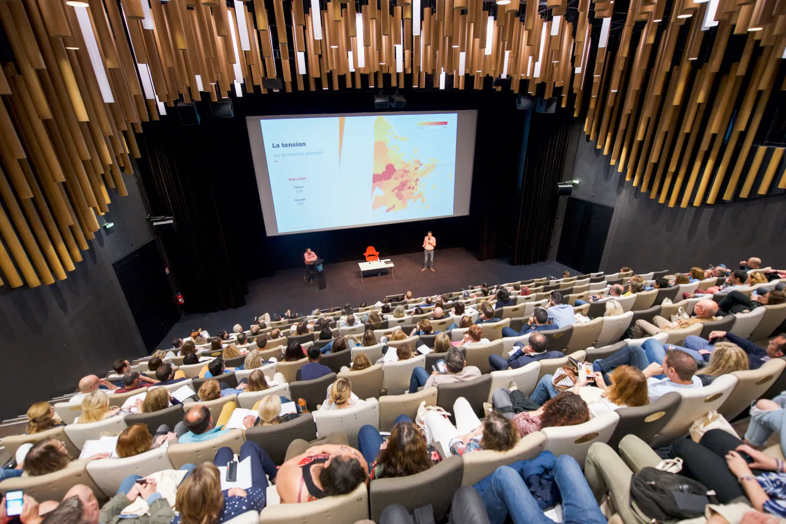 Auditorium lecture at Cité du Vin in Bordeaux showing a presentation on a large screen with a map of France, audience seated in theater-style seating, and speakers on stage with wooden acoustic panels overhead.