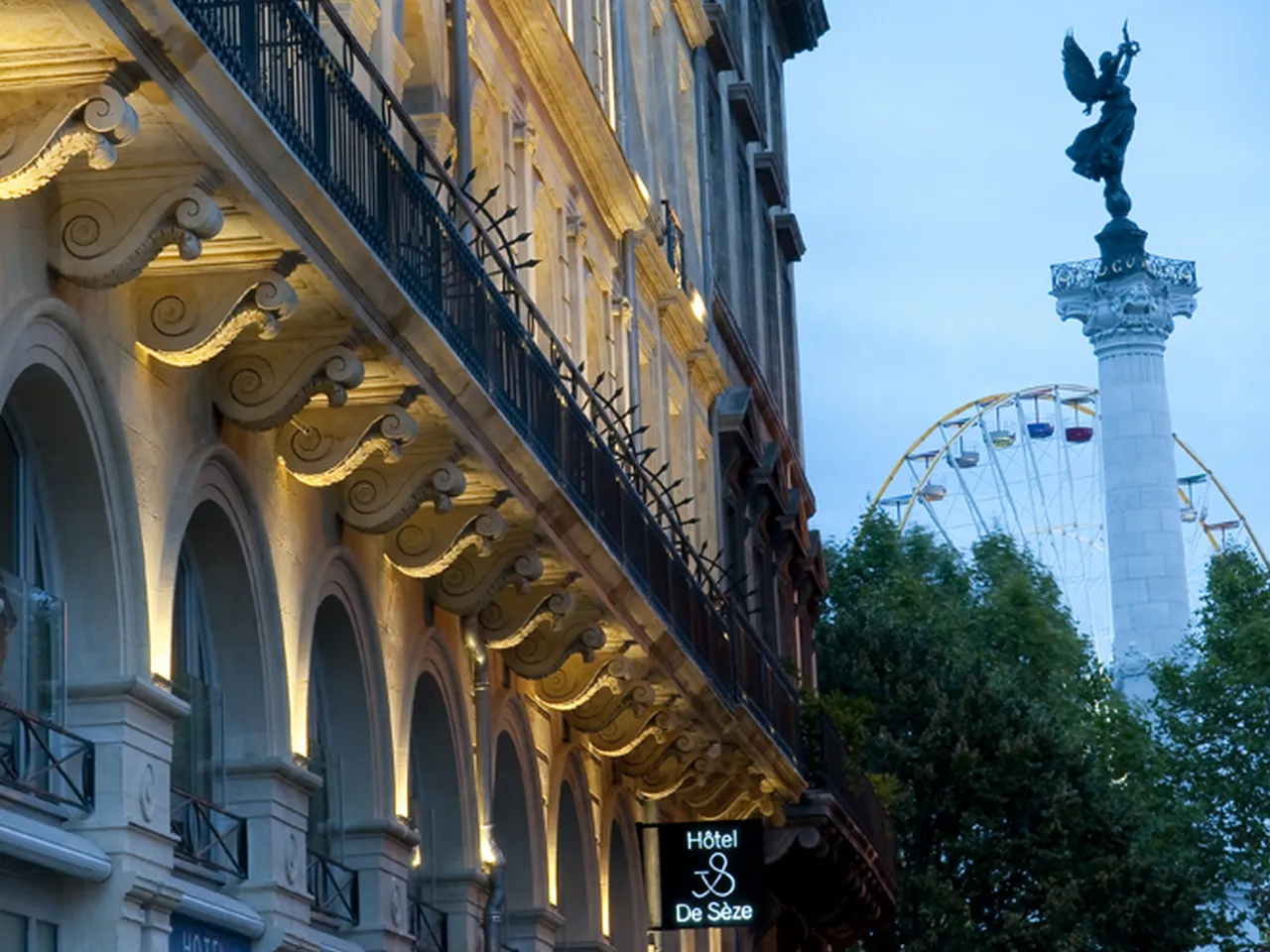 Elegant Hôtel de Sèze building in Bordeaux with ornate golden architectural details, illuminated at dusk, overlooking Trafalgar Square with its iconic column and Ferris wheel visible in the background.