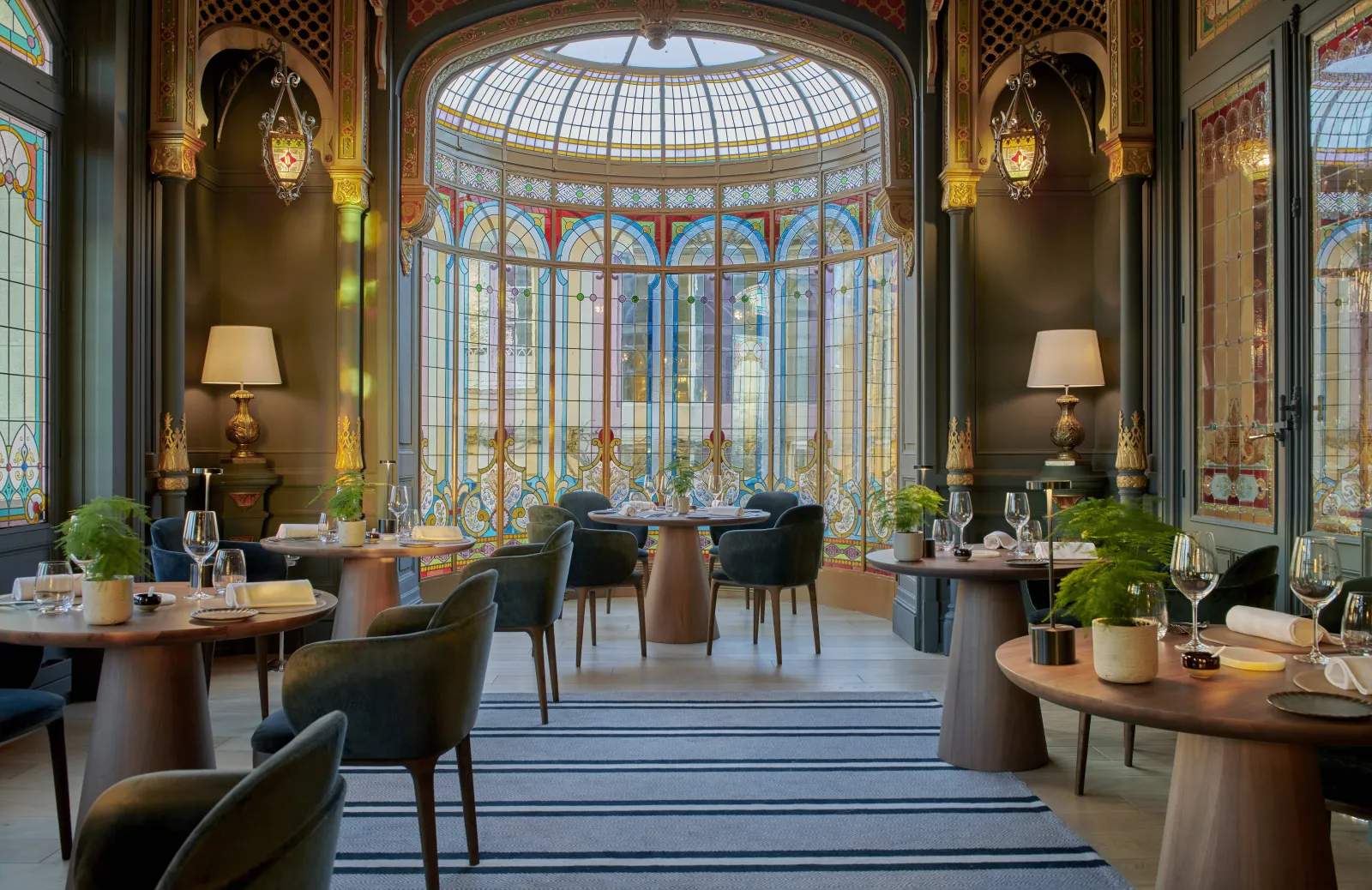 Elegant fine dining restaurant interior at Grand Barrail Château Hotel in Bordeaux, featuring round wooden tables with dark chairs, ornate stained glass windows with blue and gold patterns, decorative hanging lanterns, and a sophisticated color palette of