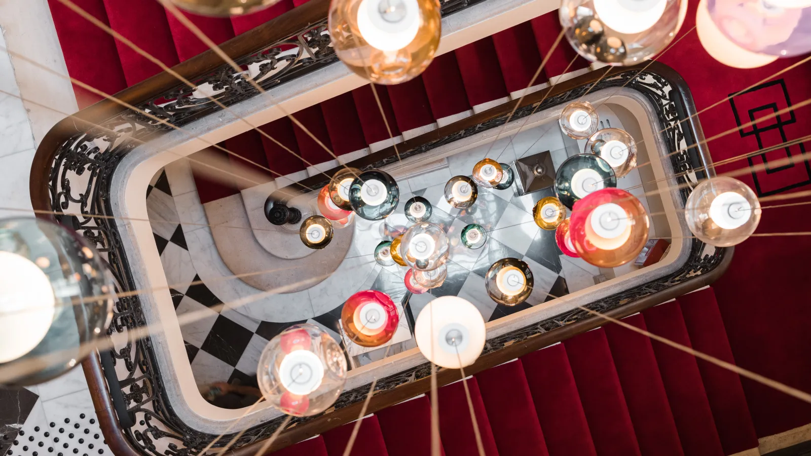 Overhead view of an ornate chandelier with multiple colored glass candle holders in amber, red, and clear tones, suspended in a stairwell with burgundy walls and decorative black railings at Hôtel Villas Foch in Bordeaux.
