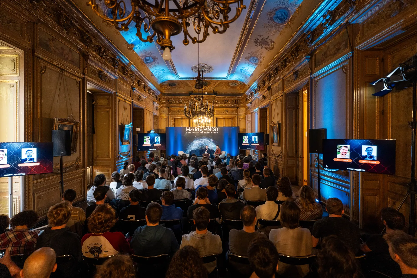 Large audience seated in an ornate classical hall with gold decorative panels and blue ambient lighting, watching a presentation on stage with the HORIZON(S) banner visible, featuring multiple screens displaying speakers.