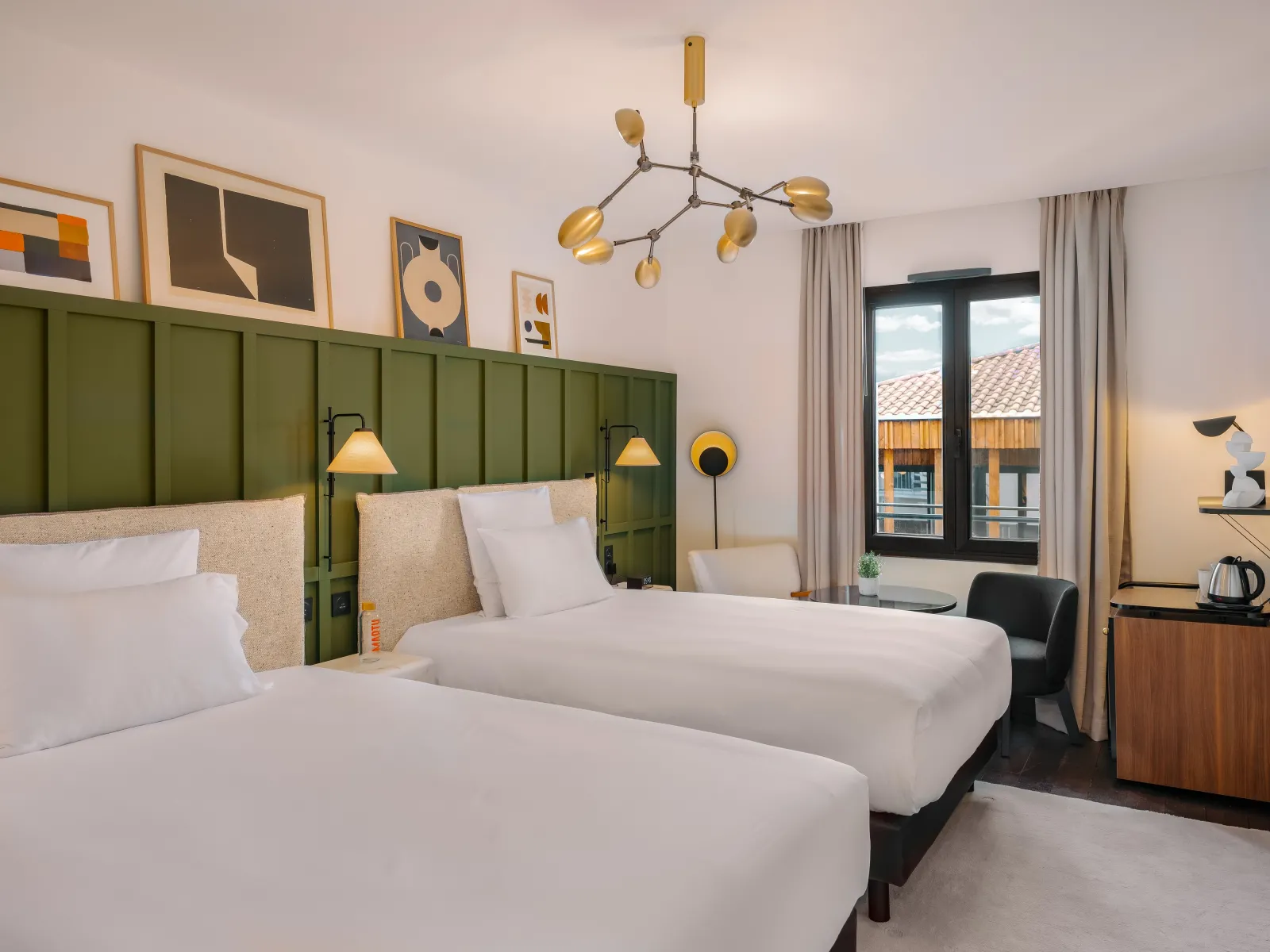 Modern hotel bedroom at Marty Hôtel Bordeaux featuring two beds with white linens, a distinctive green paneled headboard, brass pendant lighting, framed artwork, and a balcony with city views