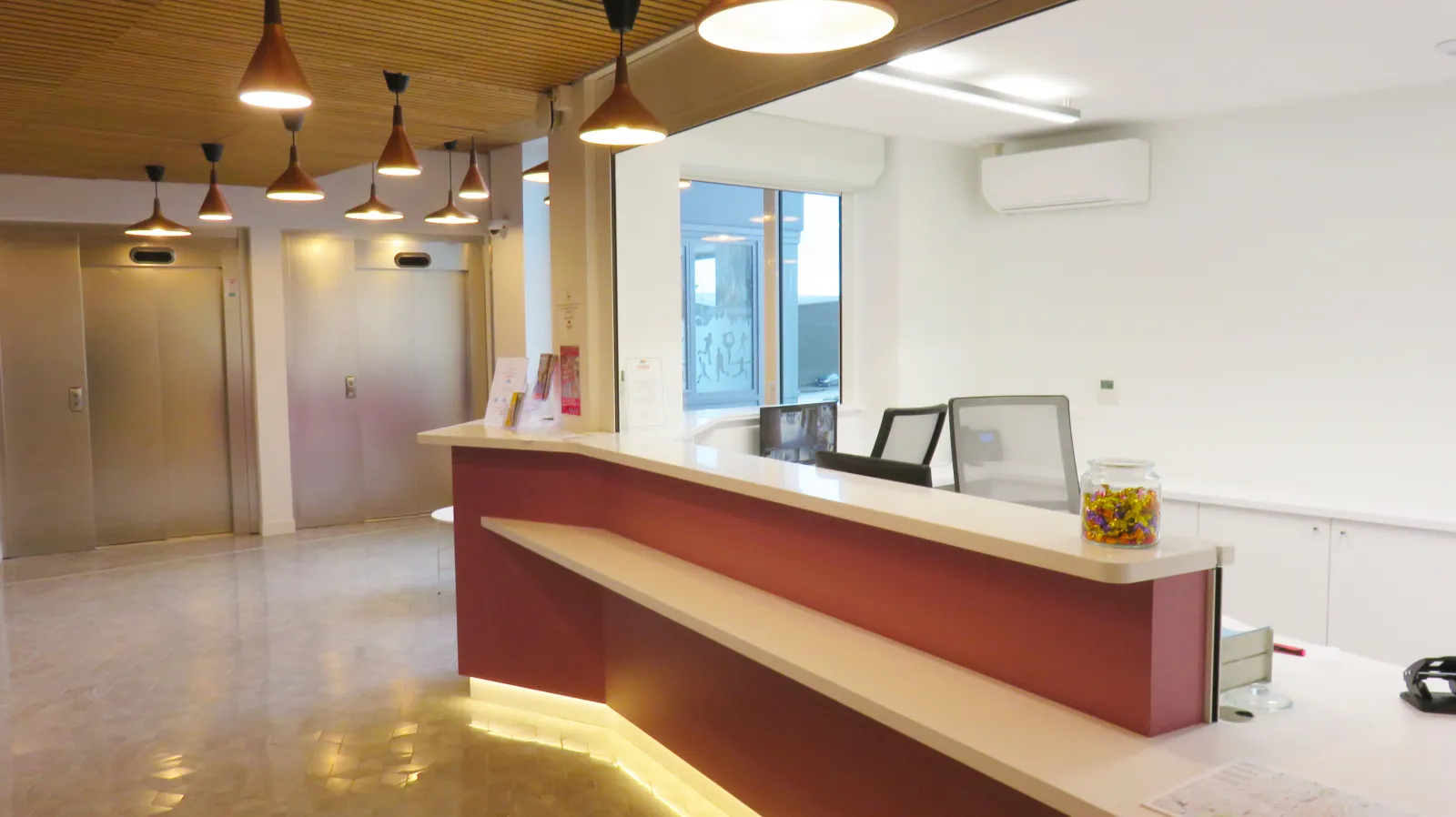 Modern reception desk in a bright lobby with red and white design, pendant lights, elevators in the background, and large windows overlooking the street