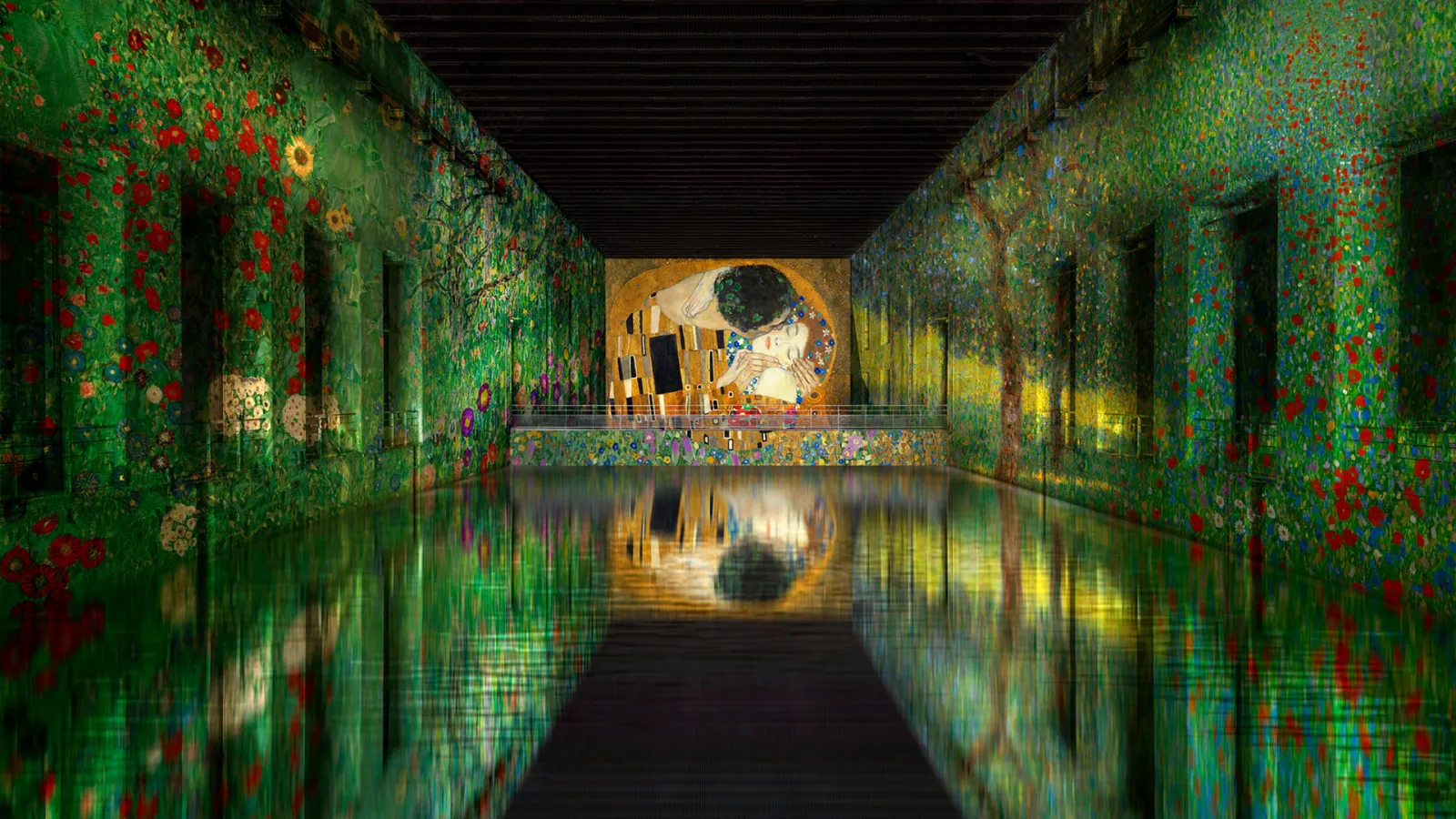 Interior of the Bassins des Lumières in Bordeaux featuring immersive digital projections on green floral-patterned walls, displaying Gustav Klimt's 'The Kiss' painting on a central screen, with reflections on the wet floor.