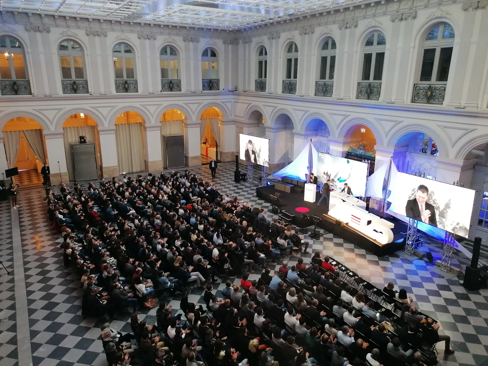 Large conference or gala event in the neoclassical Palais de la Bourse in Bordeaux, with hundreds of attendees seated facing a stage with multiple screens displaying speakers and presentations, ornate architecture with arched windows and columns visible i