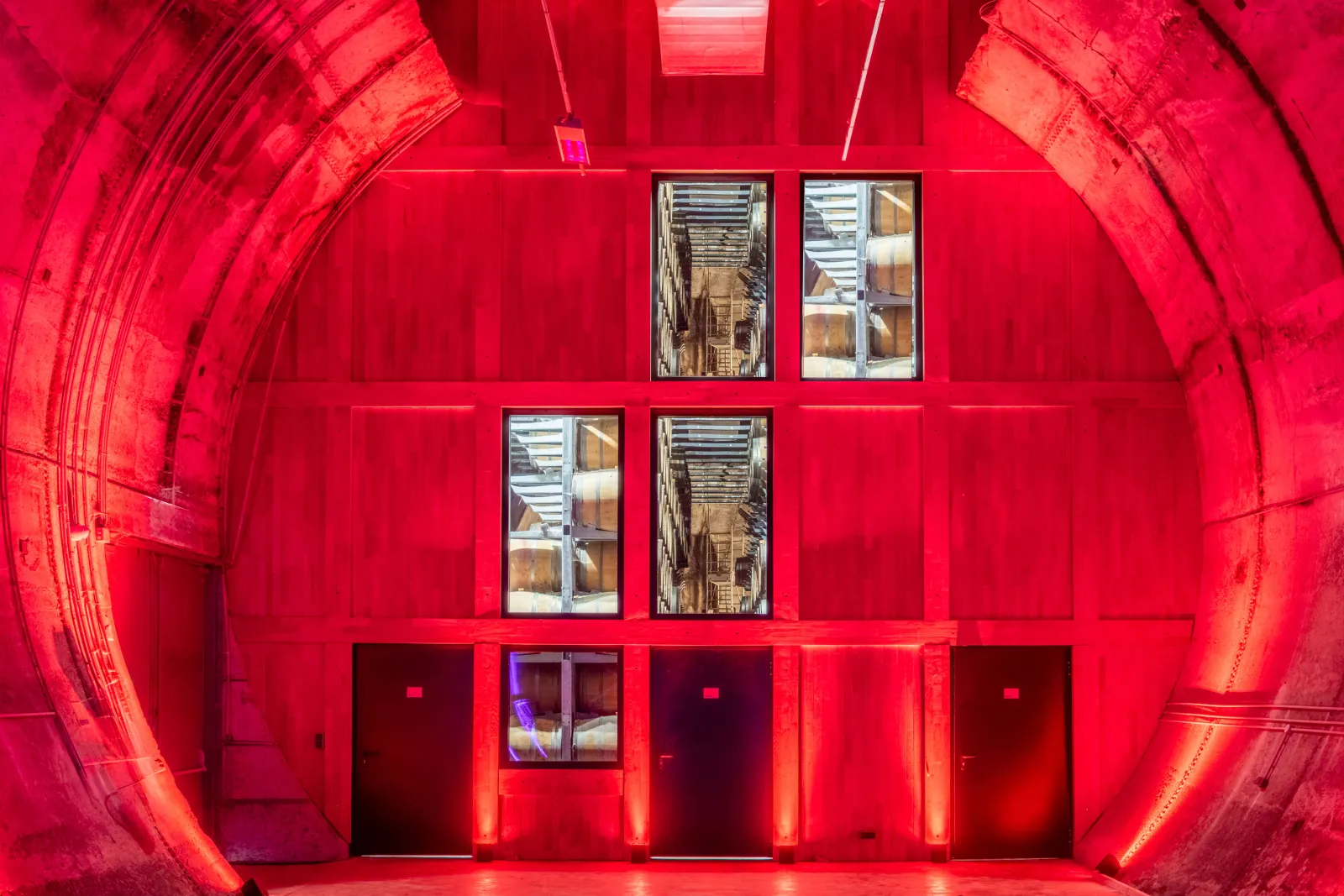 Interior of a tunnel with vibrant red neon lighting, featuring multiple windows with reflections of industrial shelving and storage areas, illuminated by bright red neon strips along the floor and architectural elements.