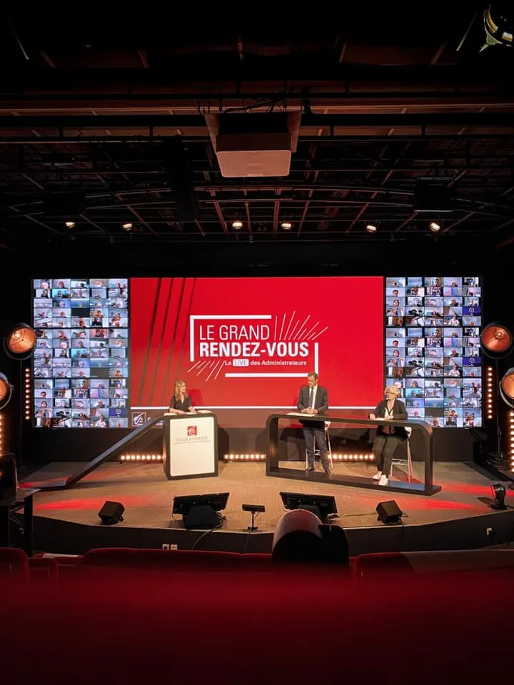 Professional conference stage with three speakers seated at a red-themed setup, large central screen displaying 'LE GRAND RENDEZ-VOUS' with grid of participant videos on side screens, modern auditorium setting with dramatic lighting.
