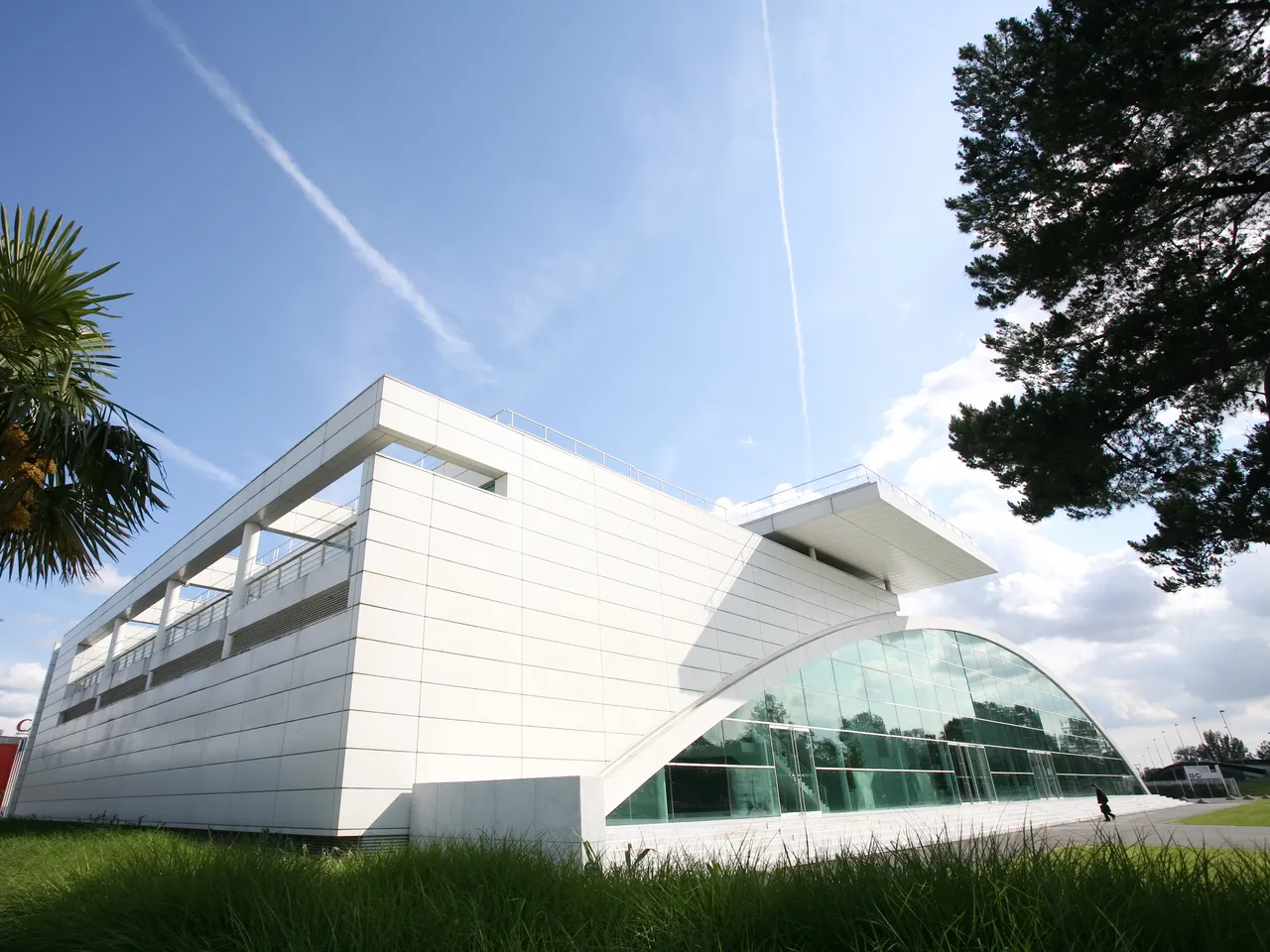 Modern white building with curved glass facade and horizontal lines, surrounded by green lawn and trees under blue sky, Bordeaux Congress Centre