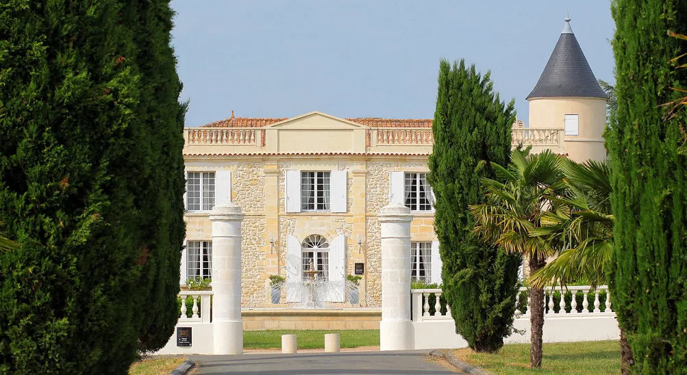 Château Lafitte in Bordeaux, France: A neoclassical mansion with cream and stone facade, white columns, mansard roof with dormer windows, round tower with conical roof, flanked by tall cypress trees and palm trees, featuring manicured gardens and balustra