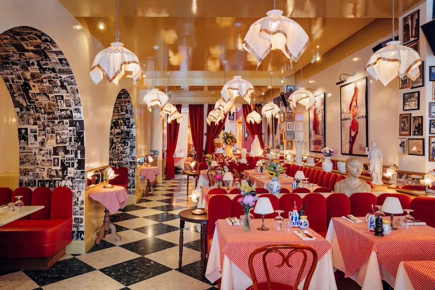 Interior of Osteria Palatino restaurant in Bordeaux featuring elegant red velvet seating, hanging sculptural lamps, checkered black and white floor, decorative archways with photographs, draped red curtains, and tables set with pink linens and floral arra