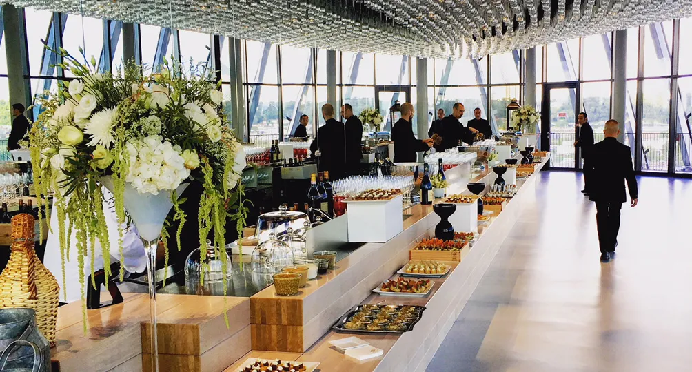 Elegant catering reception in a high-rise venue with floor-to-ceiling windows overlooking Bordeaux, featuring long service counters with white floral arrangements, appetizers, and professionally dressed staff in black attire.