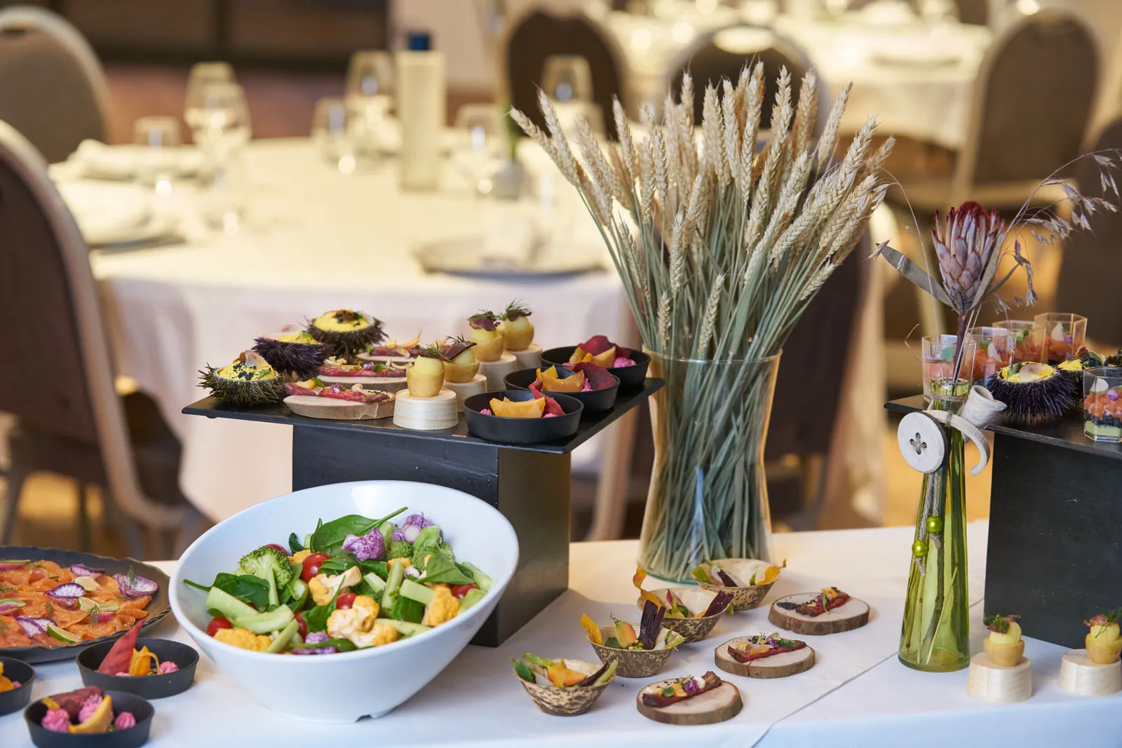 Elegant catering display at Vatel Hotel Bordeaux featuring gourmet appetizers, a fresh vegetable salad in a white bowl, wheat arrangements in glass vases, and decorative dishes with edible flowers on tables set for a formal event.