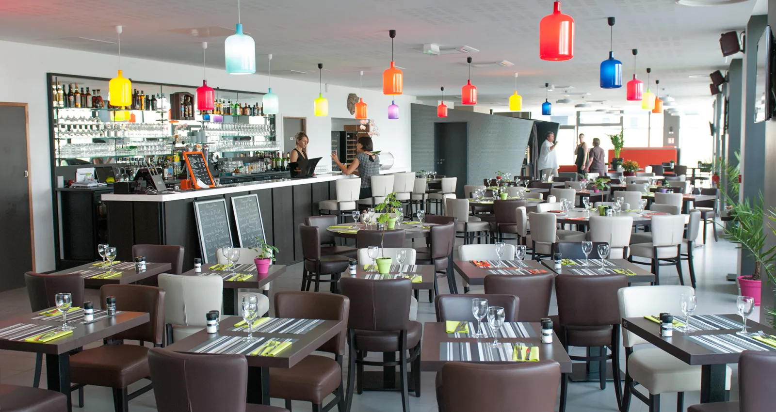 Modern restaurant interior with brown leather seating, dark tables with striped placemats, colorful hanging pendant lights in red, yellow, blue and pink, white and black bar area with bottles displayed, and plants as decoration.