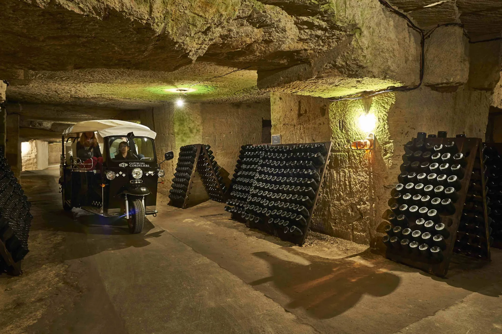 Underground wine cellar in Bordeaux's Cordeliers district featuring rows of wine bottles stored in dark wooden racks, with a small tourist tuk-tuk vehicle and atmospheric lighting illuminating the stone vault ceiling.