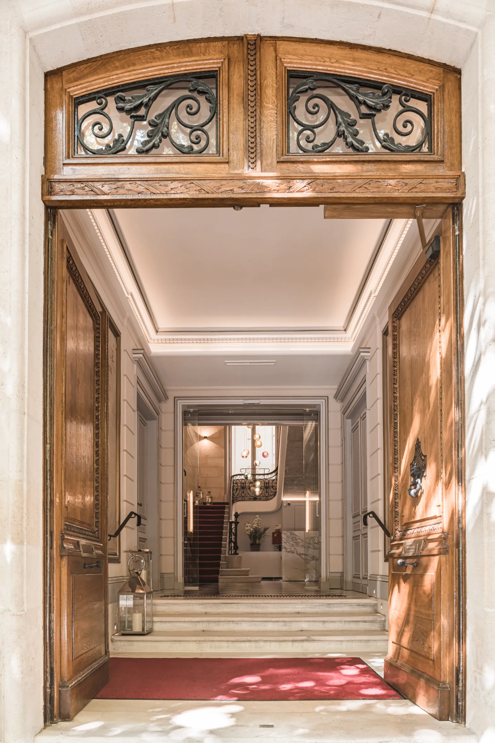 Grand wooden double doors with ornate wrought iron transom panels opening to an elegant hallway with high ceilings, classical architecture, and a red entry mat at Hôtel Villas Foch in Bordeaux.