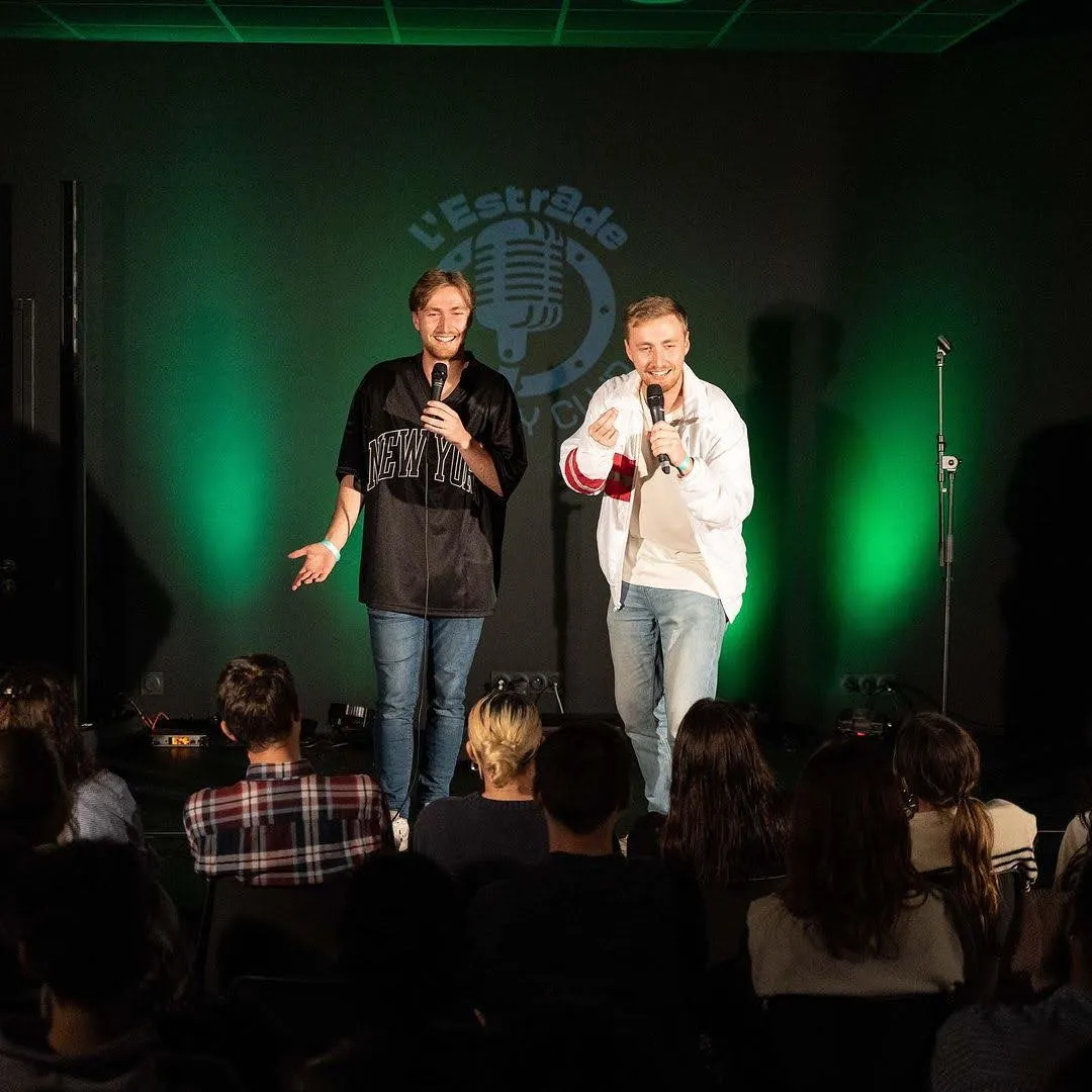 Two men performing on stage at L'Estrade in Bordeaux, holding microphones under green stage lighting, with an audience visible in the foreground.
