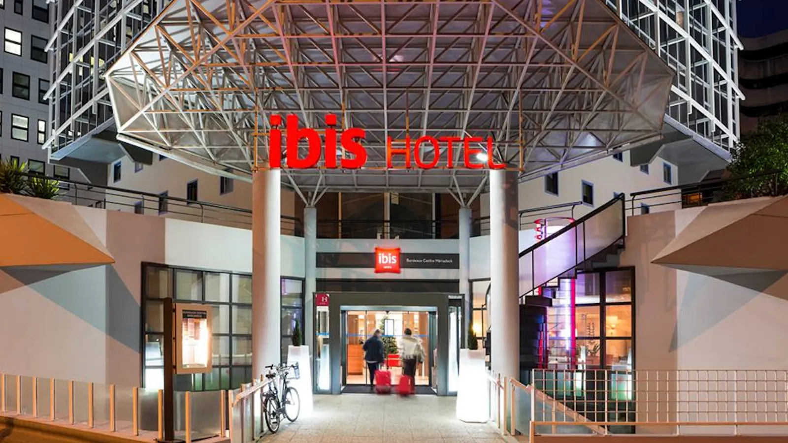 Night view of the Ibis Hotel Bordeaux Centre Mériadeck entrance, featuring a modern architectural canopy with metal framework, illuminated red signage, and contemporary urban setting with surrounding buildings.