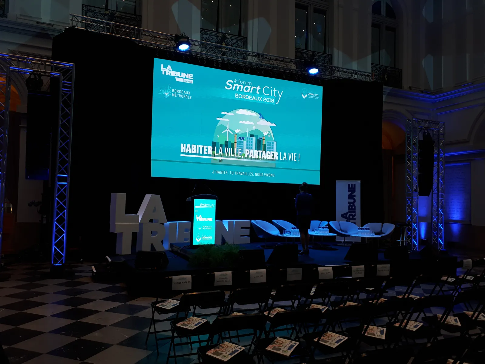 Large screen displaying the Smart City Forum Bordeaux 2018 event with turquoise branding, showing an illustrated smart city with wind turbines and the tagline 'Live the city, Share life' in an auditorium setting with audience seating and LA TRIBUNE newspa