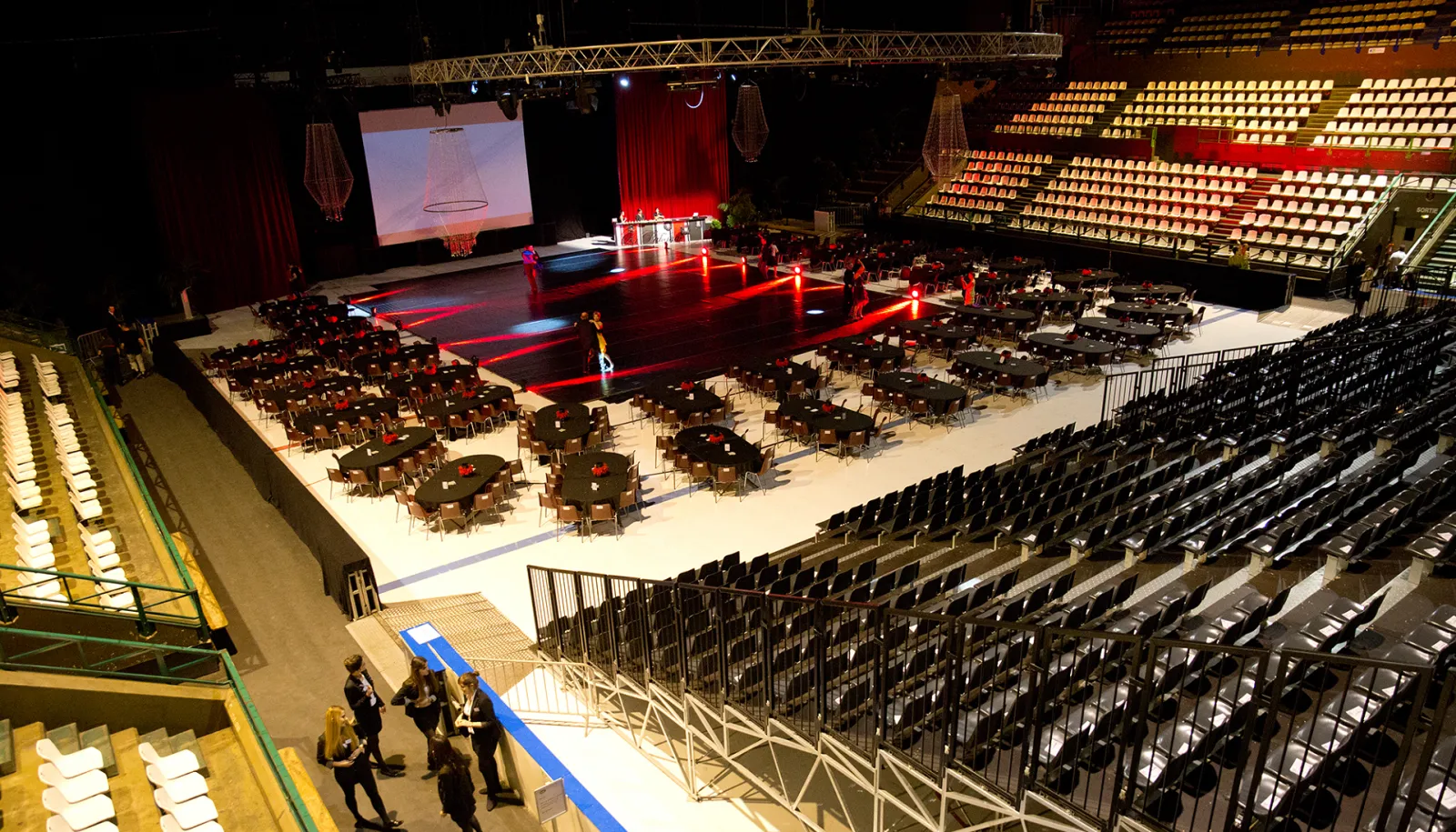 Aerial view of a large indoor arena at Mériadeck ice rink in Bordeaux, with a red-lit stage in the center, round tables with guests, theater seating, and a projection screen displaying an architectural design.