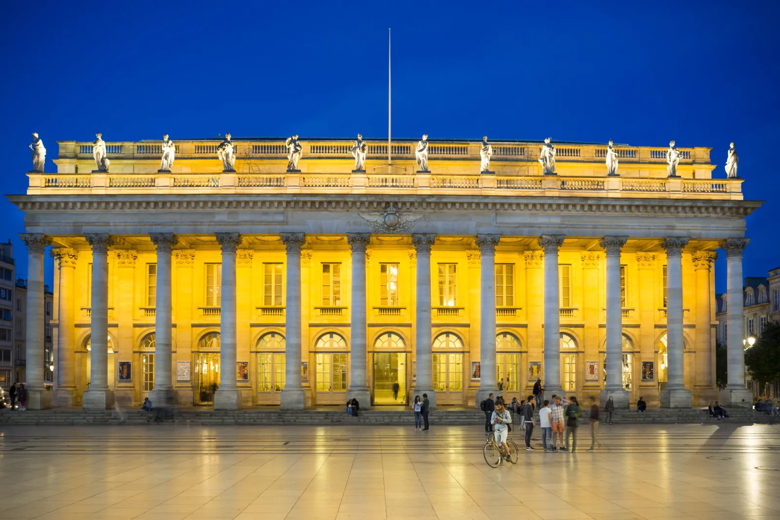 The Grand Théâtre of Bordeaux illuminated at night, featuring a classical facade with Corinthian columns, statues on the roofline, and a flagpole, with people and a cyclist in the plaza below against a deep blue evening sky.
