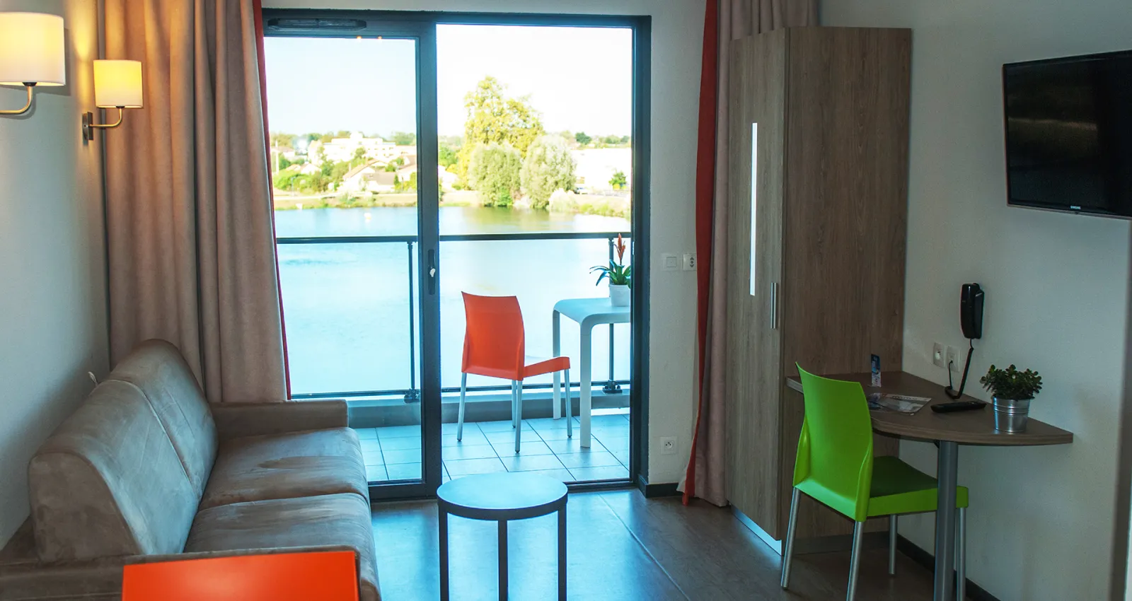 Modern hotel room with river view, featuring a gray sofa, work desk with green chair, flat-screen TV, and a balcony overlooking water and green landscapes near Bordeaux.