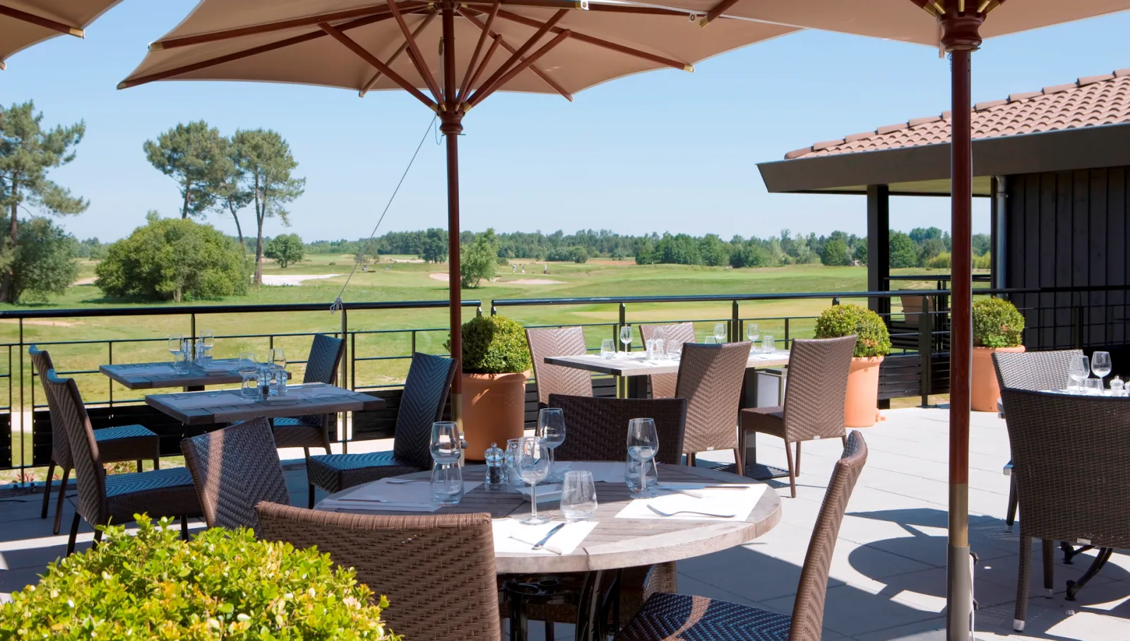Outdoor dining terrace at a luxury hotel with wicker chairs, round and rectangular tables under umbrellas, overlooking vast green vineyards and countryside landscape in Bordeaux region.