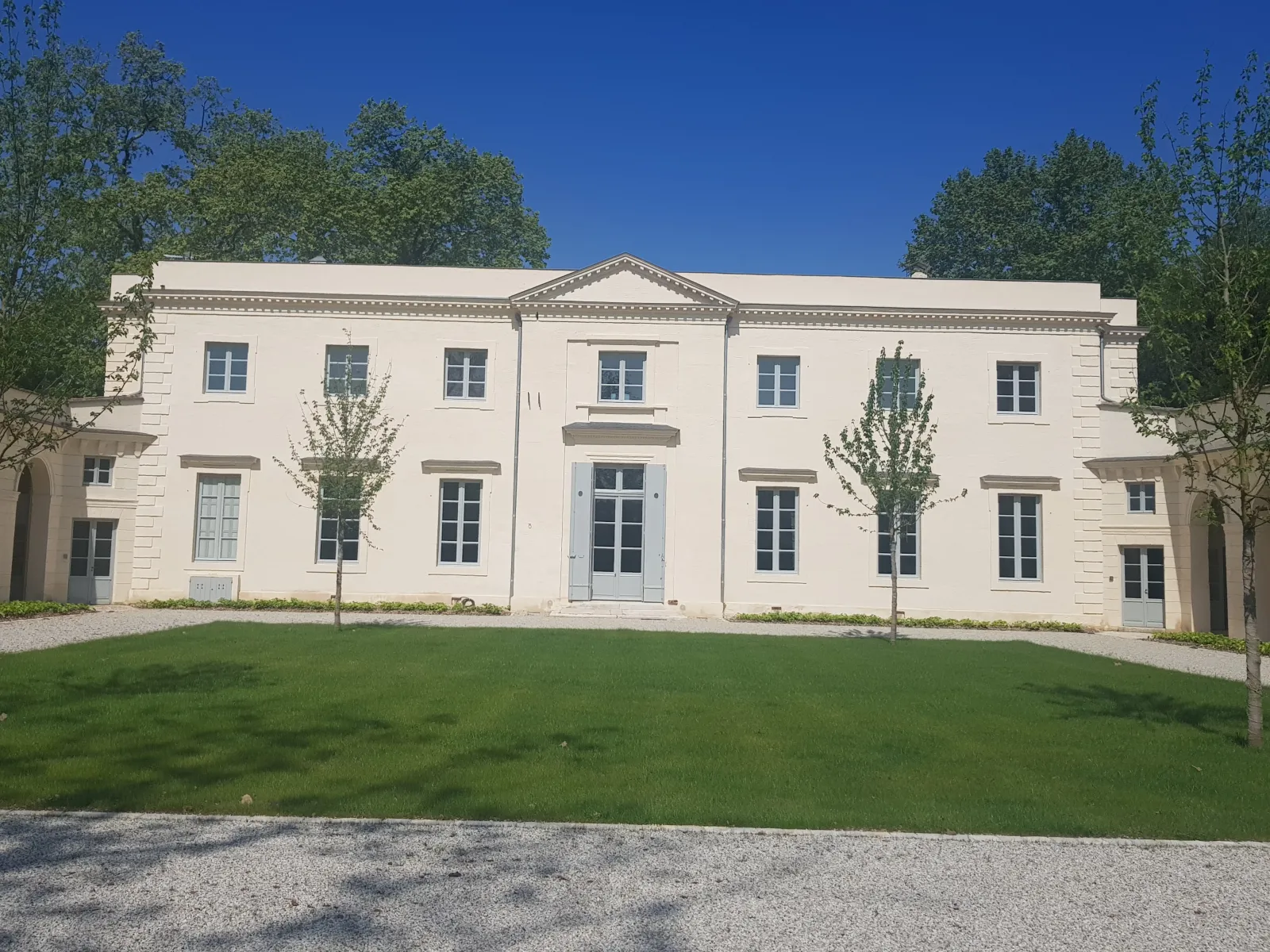 A neoclassical cream-colored manor house with symmetrical facade, multiple windows, central entrance, and triangular pediment, surrounded by manicured lawns and young trees under clear blue sky.