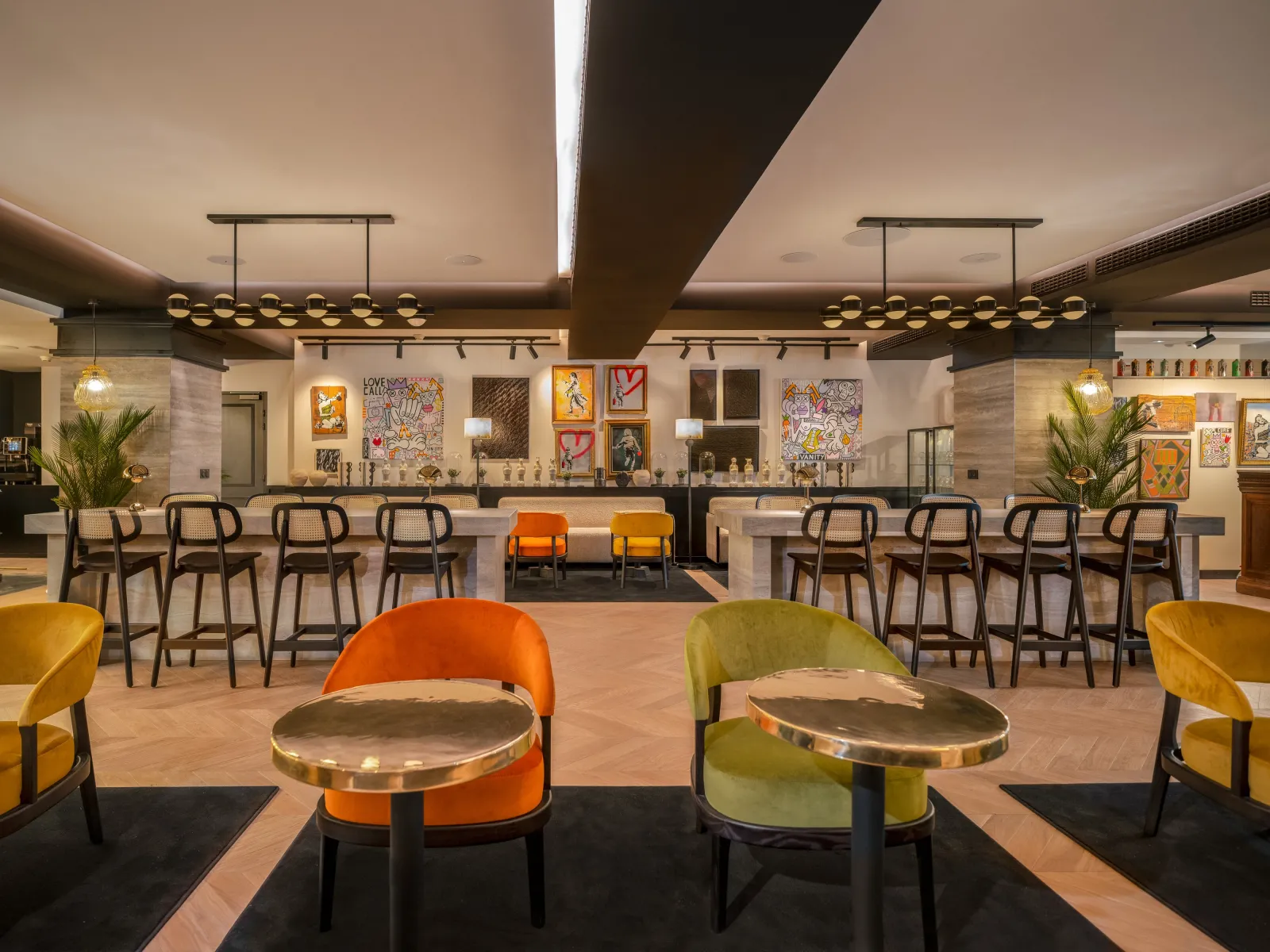 Modern hotel bar and lounge area at Marty Hôtel Bordeaux featuring colorful upholstered chairs, brass-topped tables, dual bar counters with pendant lighting, and contemporary artwork on the walls.