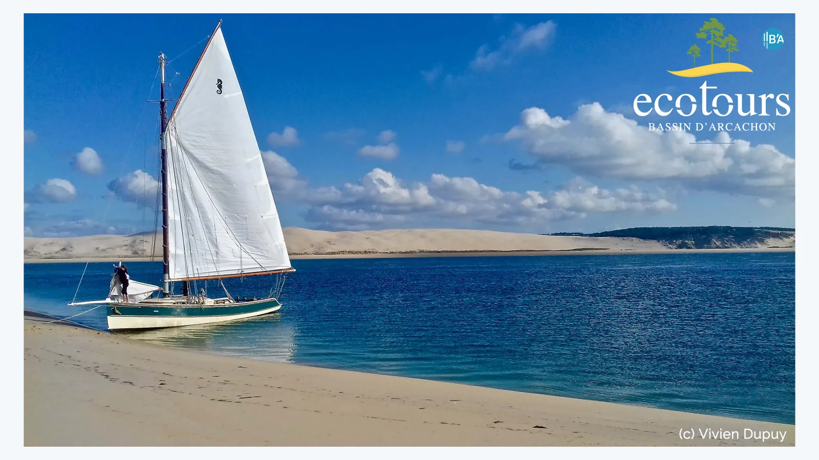 Traditional sailing boat with white sail anchored in shallow turquoise water off a sandy beach, with sand dunes visible in the background under a blue sky with white clouds.