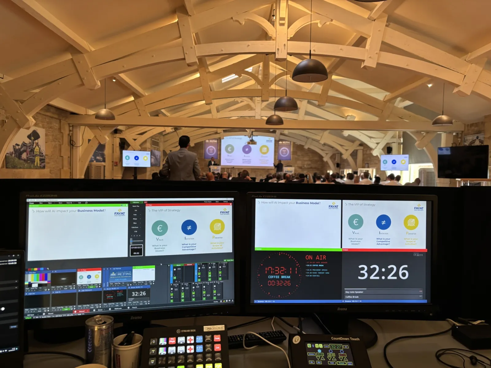 Professional broadcasting studio setup with dual monitors showing live stream controls and presentation slides, featuring a wooden-beamed event venue with audience and speakers visible on stage in the background.