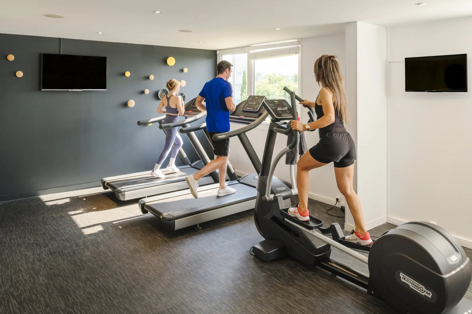 Modern home gym with three people exercising on treadmills and an elliptical machine. The bright room features gray walls with decorative gold accents, two wall-mounted TVs, large windows with natural light, and dark flooring.