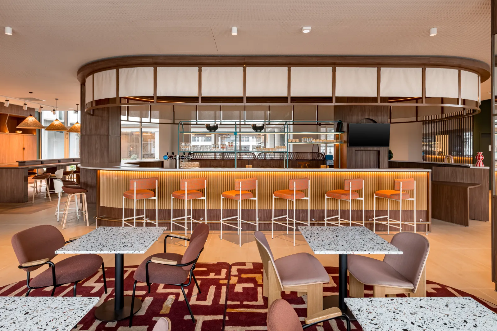 Modern hotel lobby and bar area of Hampton By Hilton Bordeaux Mérignac featuring a curved wooden counter with orange bar stools, contemporary seating areas with burgundy chairs, terrazzo tables, and floor-to-ceiling windows with natural light