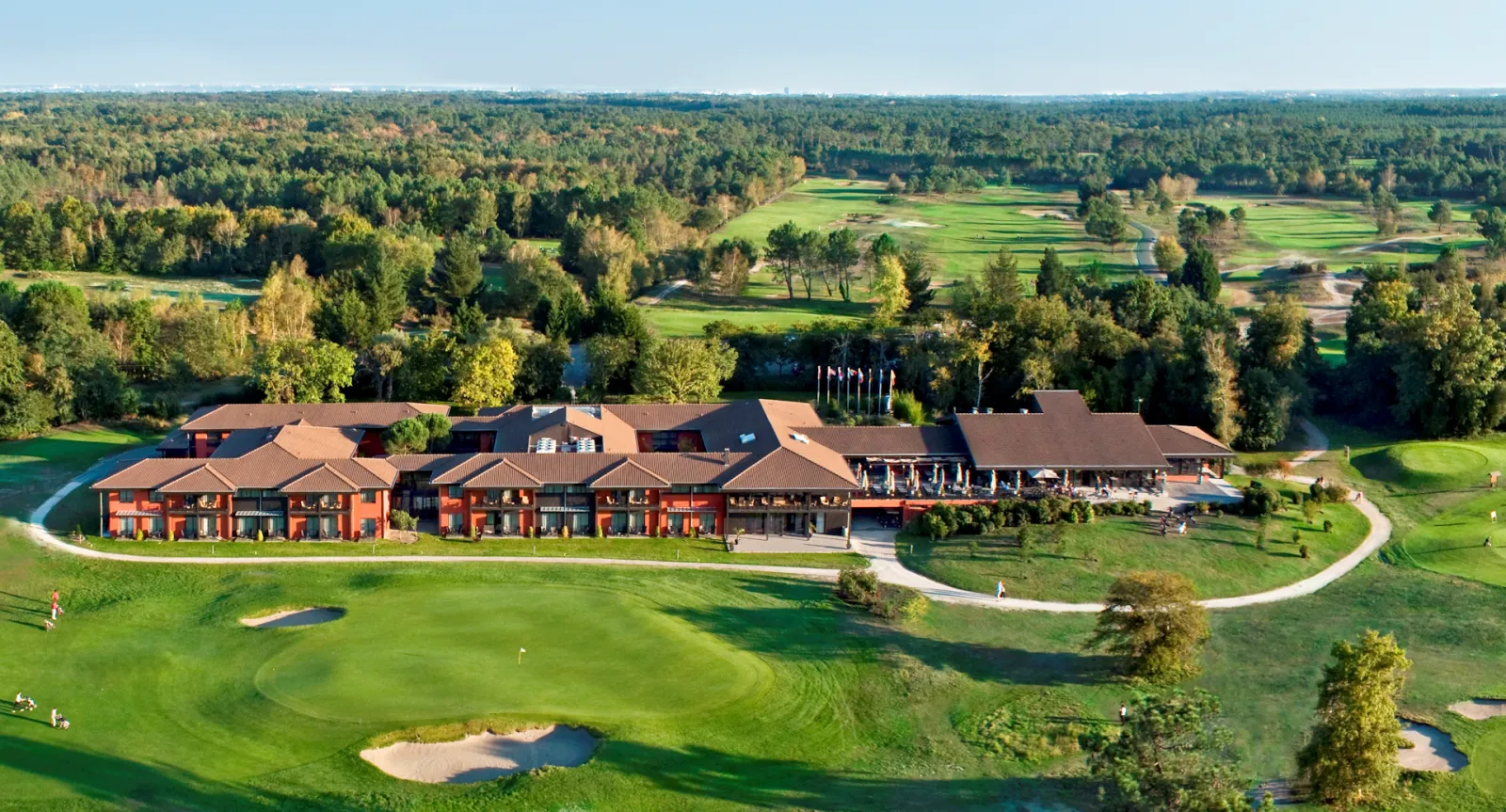 Aerial view of a luxury red-brick golf resort with modern architecture, featuring manicured greens, bunkers, and a clubhouse overlooking a river and forested landscape.