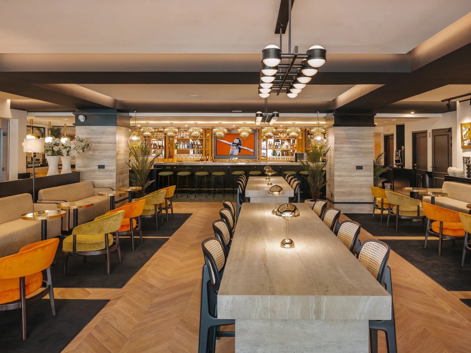 Modern hotel lounge and dining area at Marty Hotel Bordeaux featuring a long communal table, comfortable seating, bar counter with shelving, pendant lighting, and contemporary design with warm wood and neutral tones