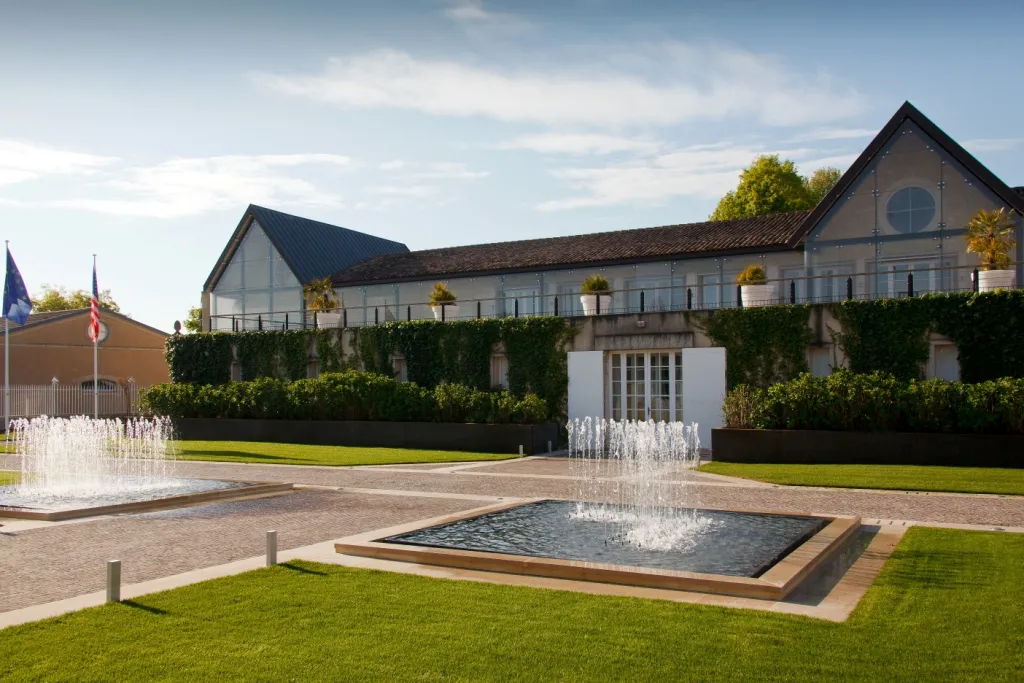 Modern architectural complex with white-walled buildings featuring dark roofs, surrounded by manicured lawns, decorative fountains, and hedges. European flags visible on the left side.