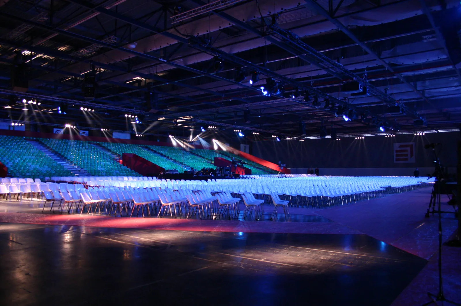 Large indoor arena at Palais 2 de l'Atlantique in Bordeaux with hundreds of white chairs arranged on the floor, illuminated by blue and red stage lighting from above, ready for an event or concert.