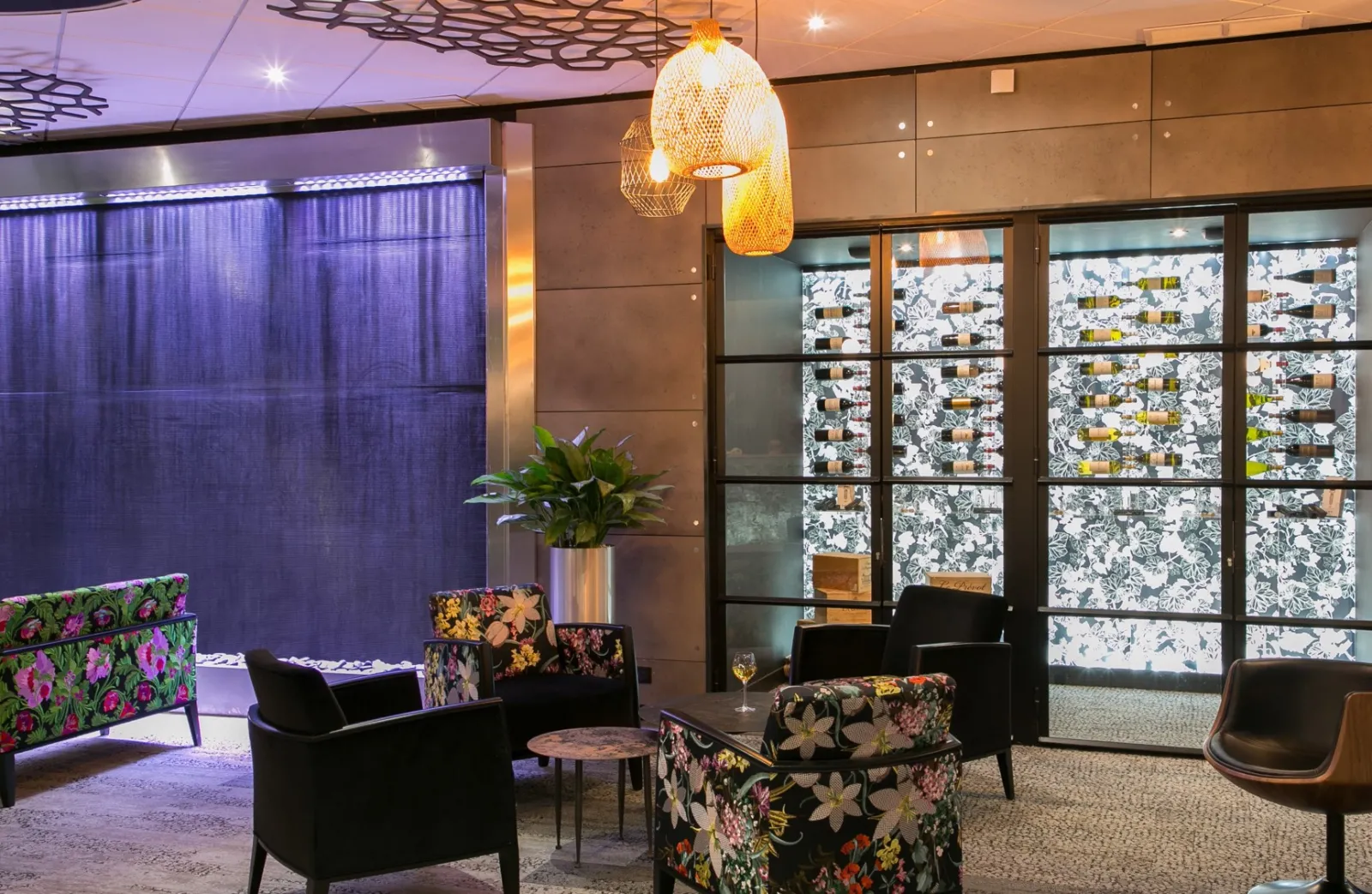 Modern hotel lobby with comfortable seating area featuring black armchairs with floral upholstered accents, decorative pendant lighting, potted plants, and large windows with patterned screens at Mercure Bordeaux Aéroport.